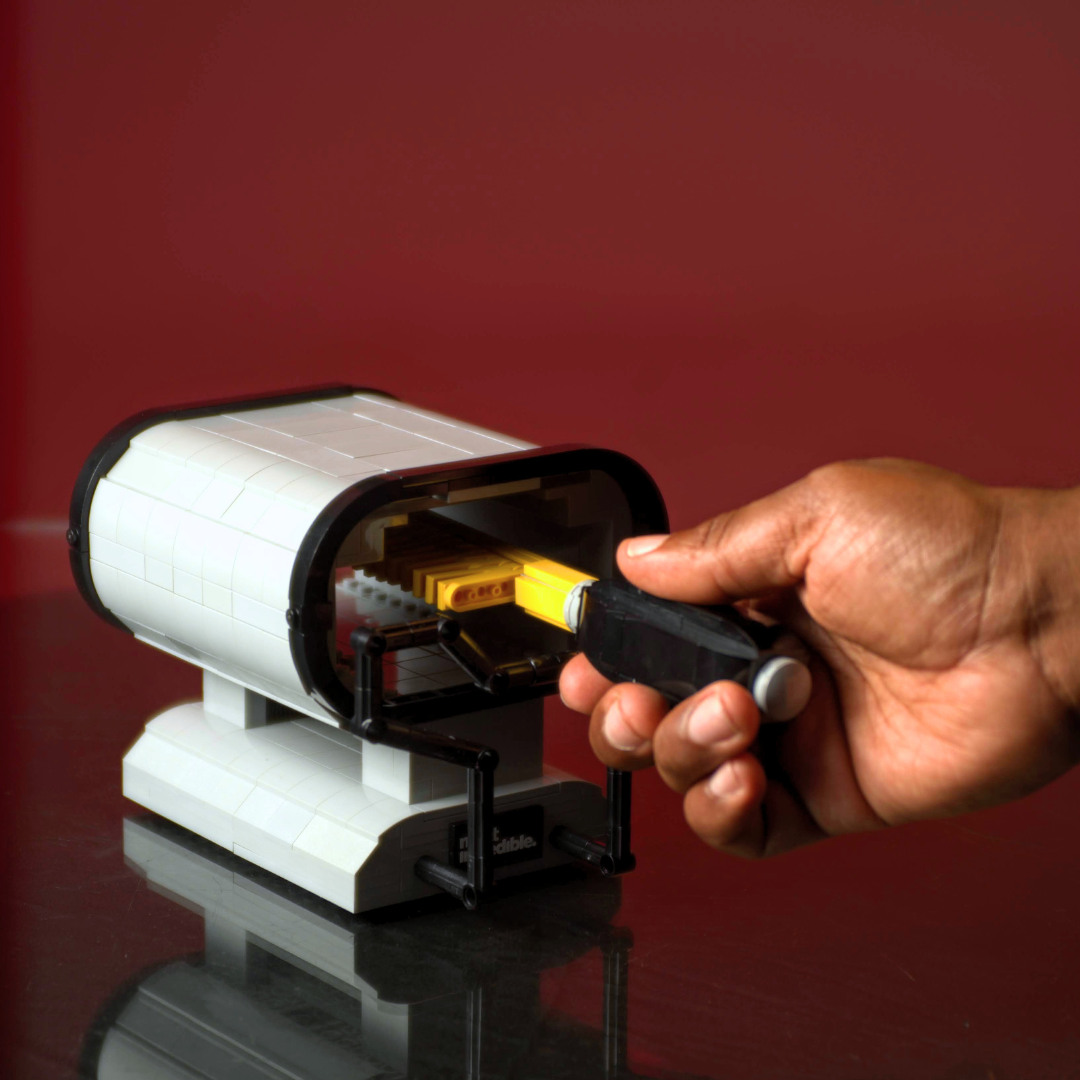 Hand inserting a yellow object into a cylindrical device on a reflective surface with a red background made of Lego